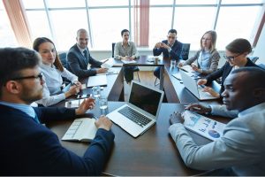 A group of people that are sat around a desk with their laptops in front of them in a meeting room setting