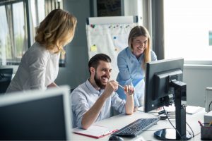A group of people that are looking over one computer. Two people are standing and one person is sitting