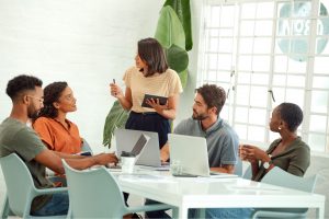 A group of people with one person standing and the others are sat in front of of their laptops
