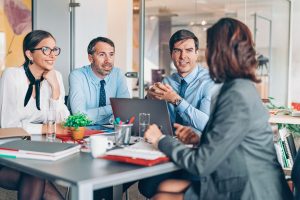 This image is a group of people who are in a office setting. They appear to be in a meeting as they have a laptop and notebooks in front of them
