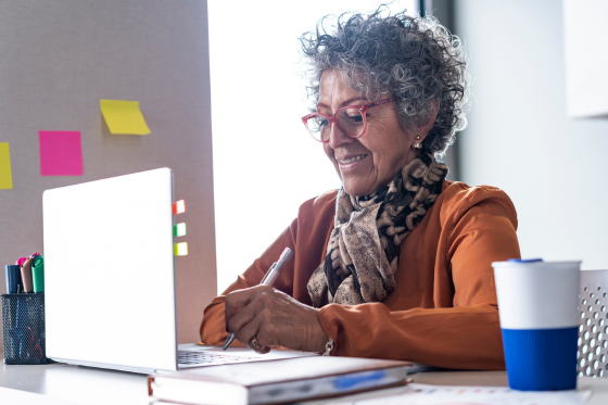 A female in an office (work from home) setting. She has a laptop in front of her and a notebook to the side of her with a cup of coffee
